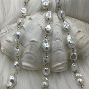 May include: Close-up of three strands of white, irregularly shaped pearls. The pearls are strung on gold-colored and silver-colored chains. The background is a white, textured surface, possibly fabric or a seashell.