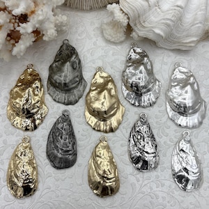 May include: A collection of ten metallic oyster shell pendants in gold, silver, and gray. Each shell has a textured surface and a small loop at the top for hanging. The pendants are arranged on a white patterned surface, with coral and shell accents.