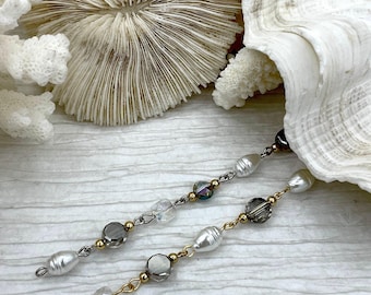 May include: Two delicate earrings with white pearls, clear and gray beads, and gold-colored accents. The earrings are displayed against a backdrop of white seashells and coral, suggesting a coastal or beach theme.