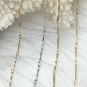 May include: Close-up of several delicate necklaces. The necklaces are gold and silver, with a bar and chain design. The background is a soft, white, textured surface. The necklaces are displayed vertically, showcasing their design.