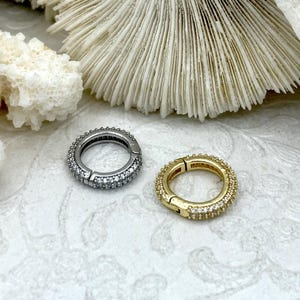 May include: Two small, round hoop earrings. One is silver-toned with clear stones, the other is gold-toned with clear stones. The earrings are set against a white background with coral.