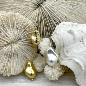 May include: Three irregular-shaped pendants in gold and silver tones. The pendants are suspended from small loops. The background features white coral and a large, textured seashell, creating a natural, organic aesthetic. Jewelry.