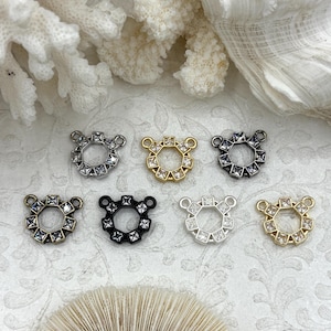 Puede incluir: Una colección de ocho pequeños dijes de joyería decorativos. Cada dije presenta un diseño circular con un patrón de gemas cuadradas y dos pequeños bucles. Los dijes están disponibles en varios acabados metálicos, incluyendo plata, oro y negro.