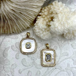 May include: Two gold-toned pendants with mother-of-pearl insets and a central clear gemstone. One pendant is square, the other rectangular. Both are surrounded by small, clear crystals. The pendants are displayed on a white, patterned surface.