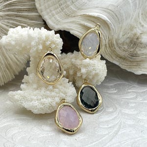 Puede incluir: Cuatro colgantes de piedras preciosas con biseles dorados. Las piedras son transparentes, blancas, rosas y gris oscuro. Los colgantes tienen forma irregular y un lazo para colgar. Se muestran sobre coral blanco.