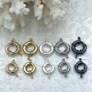Puede incluir: Una colección de cierres de joyería redondos en varios acabados metálicos, incluyendo oro, plata y negro. Los cierres están dispuestos en filas, mostrando sus diferentes colores y diseños. El fondo presenta coral blanco y una superficie estampada.