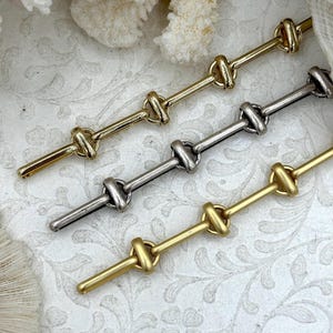 May include: Close-up of three metal bar pins in gold, silver, and brass tones. Each pin features a series of connected knot-shaped details along its length. The pins are arranged diagonally against a white floral patterned background.