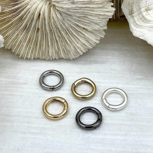 May include: Five metal spring rings in various finishes: silver, gold, gunmetal, and black. The rings are round with a hinged opening. The background features a white surface and a textured, off-white coral.