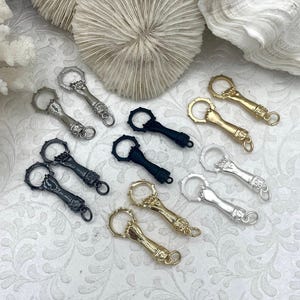 May include: A collection of bottle openers in silver, gold, and black. The openers have a hand design with a ring for leverage. The background includes white coral and a textured surface.
