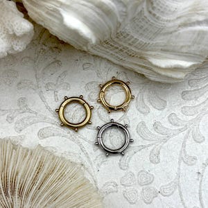 May include: Three metal hoop clasps in gold, silver, and bronze tones. Each clasp has a circular design with small, evenly spaced decorative studs around the outer edge. The clasps are displayed on a white, patterned surface with seashell accents.