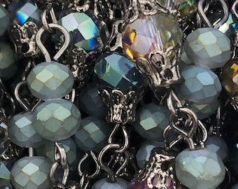 May include: Close-up of a beaded necklace or rosary. The beads are a mix of faceted, iridescent crystals and round, matte beads in shades of teal and gray. Silver-toned metal connectors and decorative caps add detail. The overall effect is intricate and elegant.