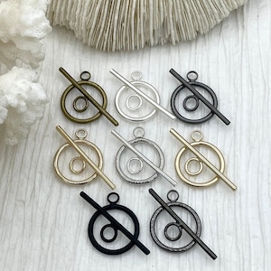 May include: A collection of toggle clasps in various metallic finishes, including gold, silver, and black. Each clasp features a circular ring with a smaller inner ring and a straight bar. The clasps are displayed on a textured, off-white surface.