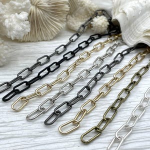 May include: Close-up of various metal chain links in black, gold, silver, and bronze colors. The chains are arranged in parallel lines, showcasing different finishes and styles. The links are oval-shaped, and the chains are displayed against a light-colored background.