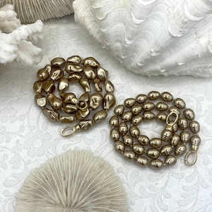 May include: Two strands of gold-colored beaded necklaces with gold clasps. The beads have an irregular, organic shape. The necklaces are displayed on a white surface with coral and seashell accents, creating a coastal theme.