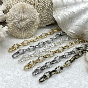 May include: Six strands of oval-link chain in various metallic finishes: gold, silver, and bronze. The chains are arranged diagonally against a backdrop of white coral and a textured, patterned surface. The chains are for jewelry making.