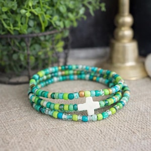 May include: A beaded wrap bracelet in shades of green, teal, and gold. The bracelet features a white cross charm. The beads are strung on a flexible cord, creating a layered effect. The bracelet is displayed on a neutral surface.