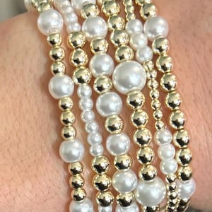 Może przedstawiać: Kolekcja bransoletek z koralików z mieszanką białych sztucznych pereł i złotych koralików. Bransoletki różnią się rozmiarem i układem koralików, tworząc warstwowy wygląd. Biżuteria jest prezentowana na nadgarstku.