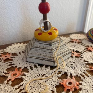 Puede incluir: Un juguete de madera vintage con una bola roja, un anillo amarillo con puntos rojos y un anillo blanco. El juguete está sobre una base piramidal blanca desgastada. Una cuerda está unida al juguete.
