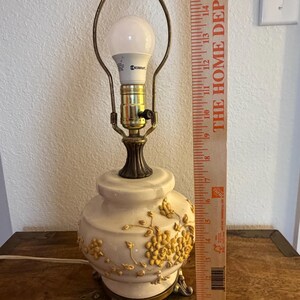 May include: A cream-colored ceramic table lamp with raised yellow floral relief patterns, a brass-toned metal base, and a harp fixture holding a white light bulb. A wooden ruler placed next to the lamp indicates a height of approximately 16 inches.