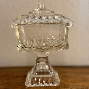 Könnte beinhalten: Eine transparente, antike Bonbonniere aus Glas mit Deckel. Die Schale ist quadratisch mit einem gewellten Rand und steht auf einem Sockel. Der Deckel hat einen dekorativen Griff. Das Glas hat ein Diamanten- und Kreismuster.