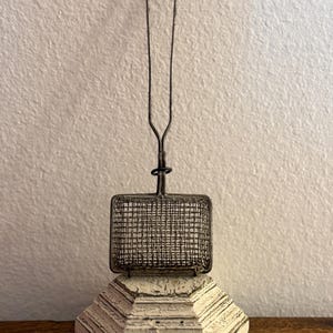 May include: A vintage metal tea strainer with a rectangular basket and long handle. The strainer sits on a white, multi-tiered, geometric base with a distressed finish. The background is a plain, off-white wall.