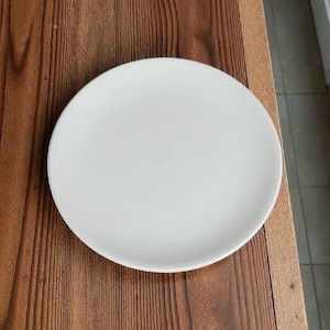 Könnte beinhalten: Ein schlichter, weißer Keramikteller, rund in der Form, liegt auf einer Holzoberfläche. Der Teller hat eine glatte, matte Oberfläche und ein schlichtes Design. Ideal zum Servieren von Mahlzeiten oder als Dekoration.