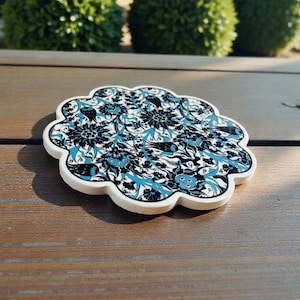 May include: A decorative coaster with a white scalloped edge. The coaster features a floral design in black and teal blue. The coaster is on a wooden surface.