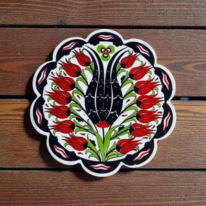 May include: A round, decorative ceramic tile with a floral design. The design features red tulips, green leaves, and a black and white border. The tile is placed on a wooden surface.