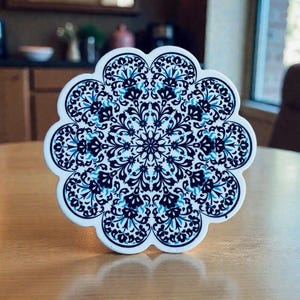 May include: A white, scalloped-edge coaster with a detailed blue and navy floral pattern. The coaster has a circular design with a symmetrical arrangement of stylized flowers and leaves. The coaster is sitting on a wooden surface.