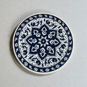 Pode incluir: Base para copos redonda com um design floral azul marinho e branco. A base apresenta um padrão central em forma de estrela rodeado por intrincados motivos florais e de folhas. A borda é adornada com pequenas flores brancas.