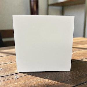 May include: A square, white planter box sits on a weathered wooden table. The planter has a clean, minimalist design, with a simple, geometric shape. The background includes a dark brown bottle and a shelf with other items.