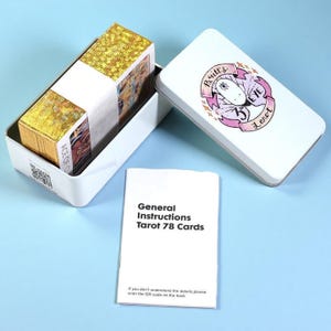 May include: A white metal tarot card box with a lid featuring a pink and purple illustration and the text "Britt's Eye Tarot." Inside, a deck of cards is visible, along with a booklet that reads "General Instructions Tarot 78 Cards."