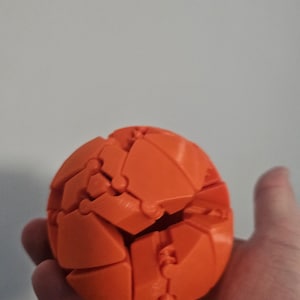 May include: An orange, spherical puzzle toy. The ball is composed of interlocking geometric shapes, creating a complex, three-dimensional design. The toy is held in a hand against a plain background. The puzzle is likely 3 inches in diameter.