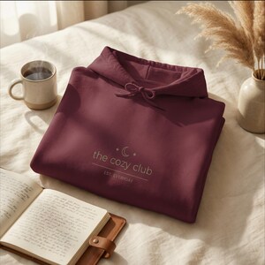 May include: A burgundy hoodie with a drawstring hood, featuring the text "the cozy club" and a crescent moon and star design. A mug of coffee and an open notebook are also visible.