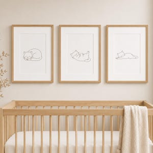 May include: Three vertical wall art prints in light wood frames, each featuring a simple black line drawing of a cat in a different sleeping position on a white background, displayed above a light wood crib with a cream-colored blanket.