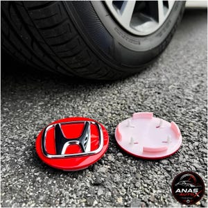 May include: Two circular Honda wheel center caps, one showing the glossy red front with a chrome H logo, and the other showing the white plastic back with four mounting clips. The caps are positioned on asphalt near a car tire.