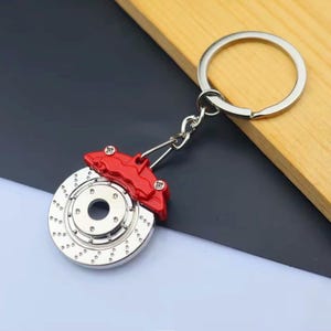 May include: A silver-colored keychain featuring a miniature car brake rotor and red caliper. The rotor has drilled holes and a central hub. The keychain is attached to a silver ring.