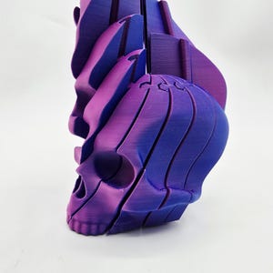 Puede incluir: Una escultura de calavera impresa en 3D en tonos morados y azules. La calavera está segmentada, con cada sección ligeramente desplazada, creando un efecto en capas. El diseño tiene un acabado liso y mate y está sobre un fondo blanco.