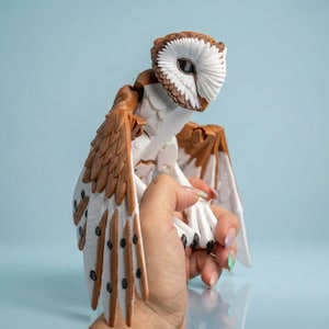 May include: A detailed owl figurine in brown and white. The owl has outstretched wings with brown and white feathers, and a white face with a brown outline. The figurine is held in a hand against a light blue background.
