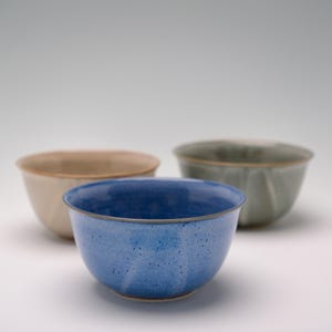 May include: Three ceramic bowls in varying colors: a speckled blue bowl in the foreground, a beige bowl, and a green bowl in the background. The bowls have a simple, rounded design.
