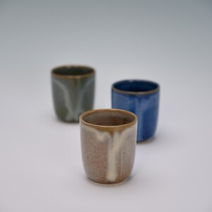 May include: Three ceramic cups in shades of brown, blue, and green. The cups have a smooth, glazed finish with a slightly irregular shape. The rims are a darker shade than the body of the cups.