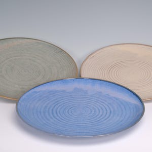 May include: Three ceramic oval plates in shades of blue, green, and beige. Each plate features a spiral design in the center. The plates are arranged on a white surface, showcasing their colors and textures. Ideal for serving food.