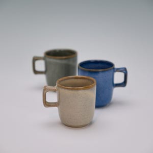 May include: Three ceramic mugs in a neutral setting. The mugs are in shades of beige, blue, and green, each with a square handle and a contrasting rim. The mugs are likely for coffee or tea.