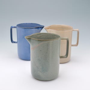 May include: Three ceramic pitchers in shades of blue, green, and beige. Each pitcher has a handle and a spout for pouring. The pitchers are arranged on a white surface.