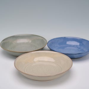 May include: Three ceramic bowls in a studio setting. The bowls are in shades of blue, beige, and green. Each bowl has a spiral pattern in the center. The bowls are shallow and have a simple, modern design.