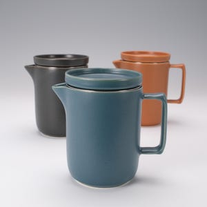 May include: Three ceramic mugs with lids and handles, arranged in a group. The front mug is teal, the back left mug is charcoal gray, and the back right mug is terracotta orange. Each mug features a cylindrical body, a spout, and a flat lid.