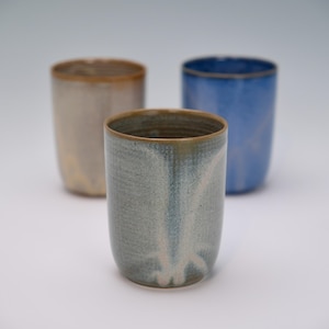 May include: Three ceramic tumblers in shades of blue, gray, and beige. Each tumbler has a rounded base and a slightly flared rim with a brown accent. The tumblers are arranged on a white surface.