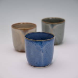 May include: Three ceramic cups in a studio setting. The front cup is a speckled blue with a brown rim. The other two cups are out of focus, one is beige and the other is gray. The cups are cylindrical and have a smooth finish.