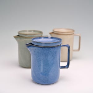 May include: Three ceramic pitchers with lids. The front pitcher is a speckled blue, with a handle and a spout. The other two pitchers are in the background, one is a muted green and the other is a cream color. All have a similar design.