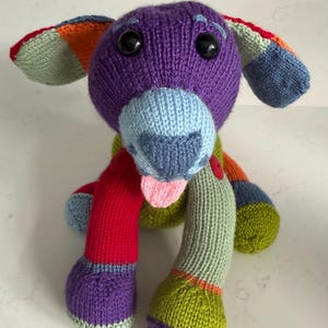 May include: A multi-coloured knitted stuffed animal shaped like a dog. The toy features a purple head, light blue muzzle, and a small pink tongue. Its body and limbs are constructed from various knitted patches in red, green, orange, and blue.